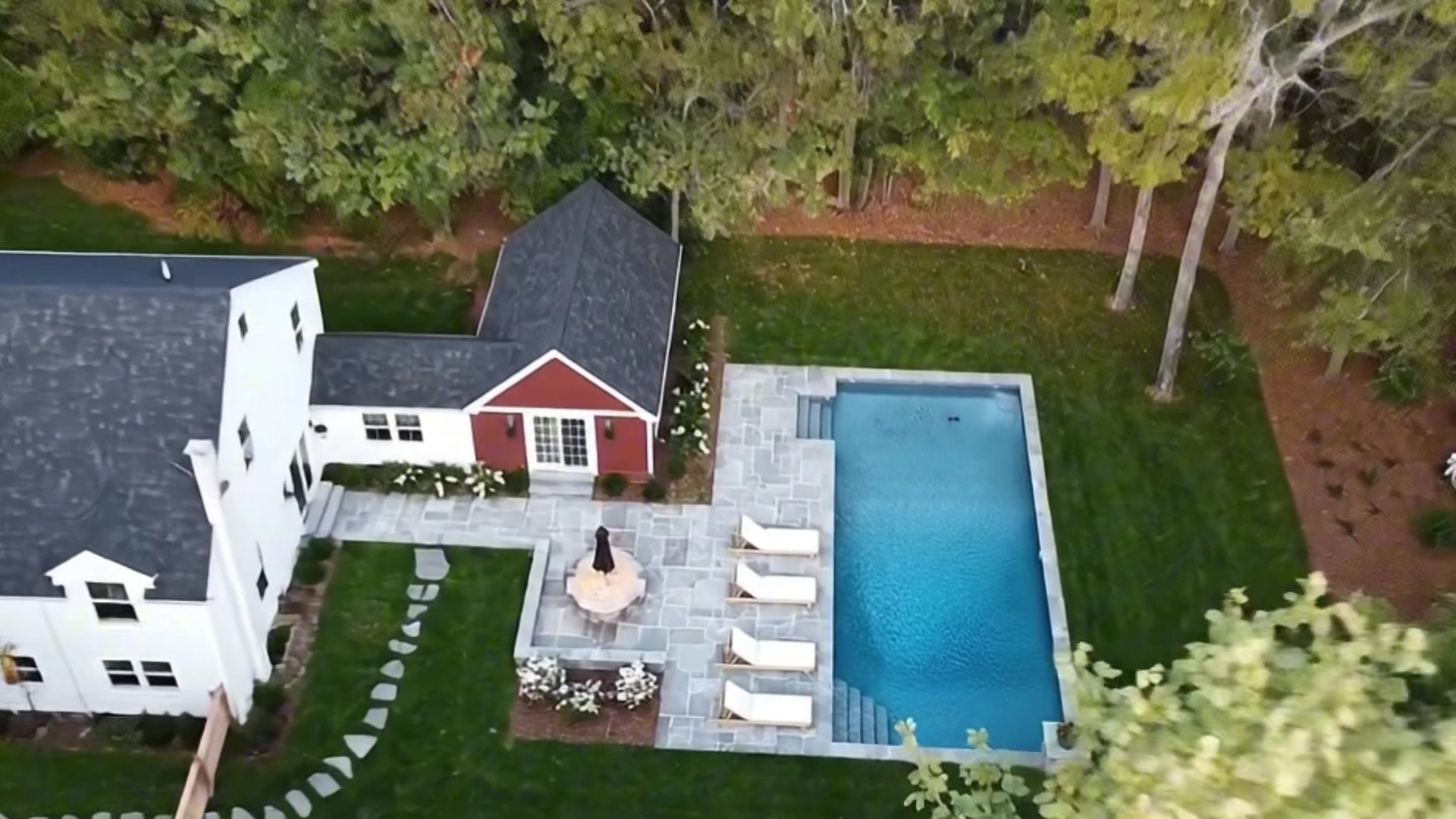 Aerial view of the property showing the white main house, red pool house, stone patio with loungers, and in-ground swimming pool surrounded by lush green lawn and trees