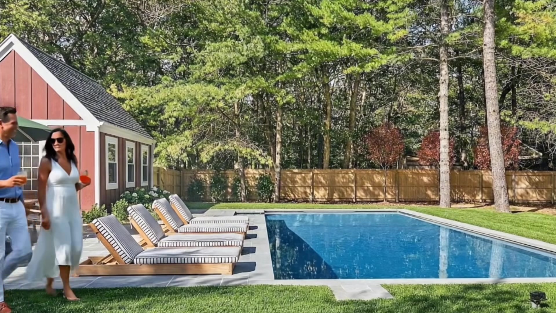 Ground-level view of the heated pool with striped cushion loungers on the stone patio, red pool house with white trim, and tall trees providing a private wooded backdrop