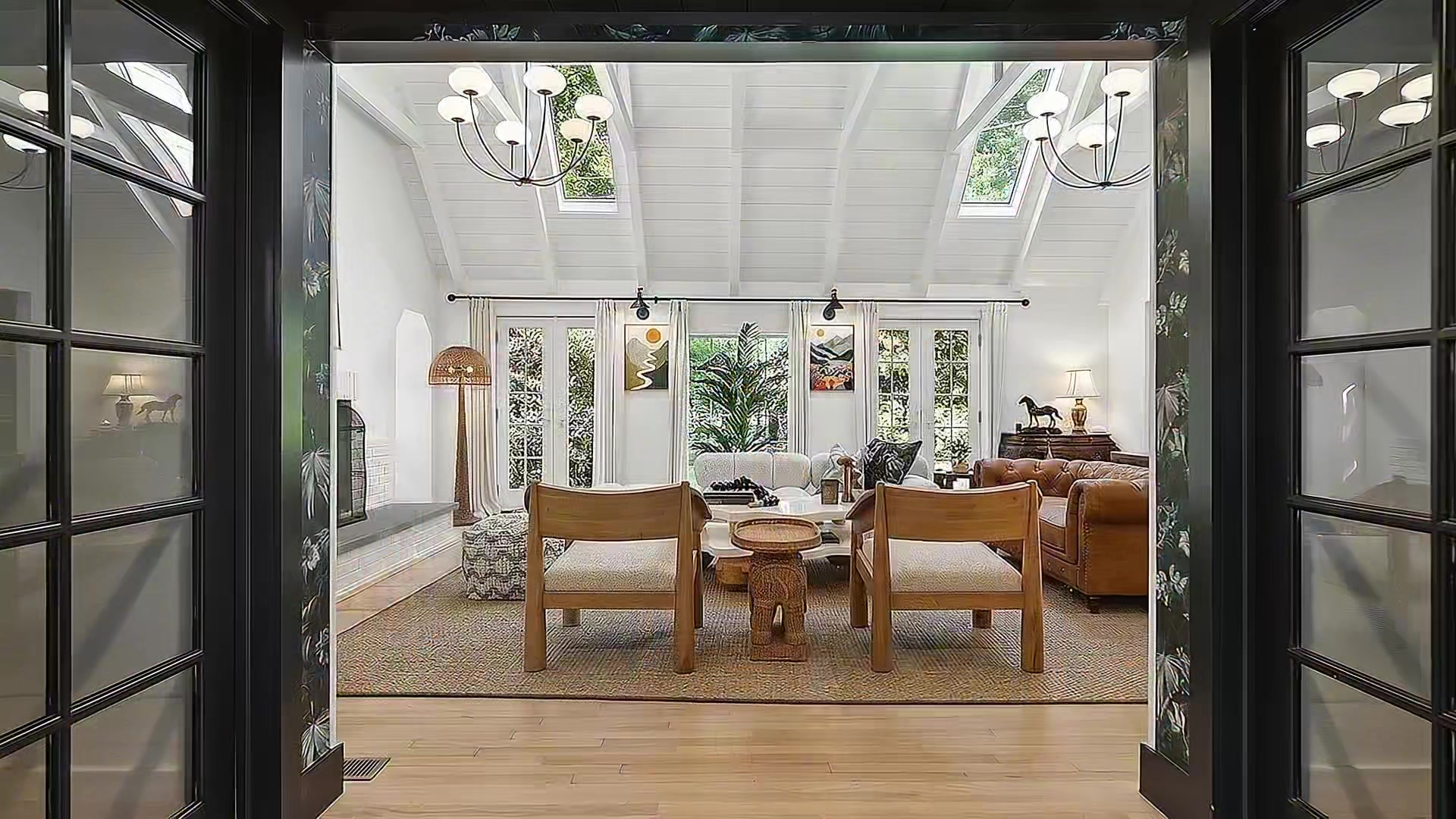 View through open black French doors into the vaulted-ceiling living room with skylights, chandeliers, mid-century furniture, and greenery visible through the far windows