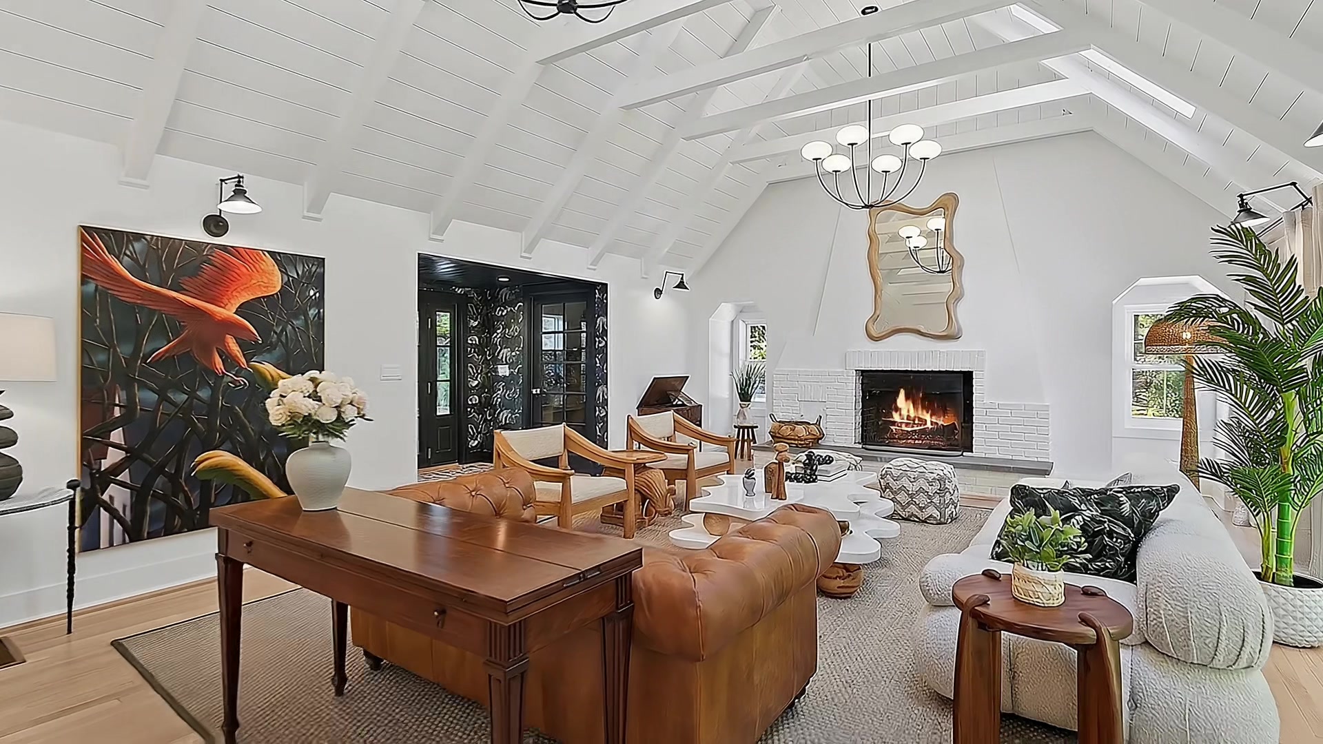 Spacious living room with dramatic vaulted shiplap ceiling, wood-burning brick fireplace, tufted leather Chesterfield sofa, mid-century wooden chairs, and a large tropical bird painting