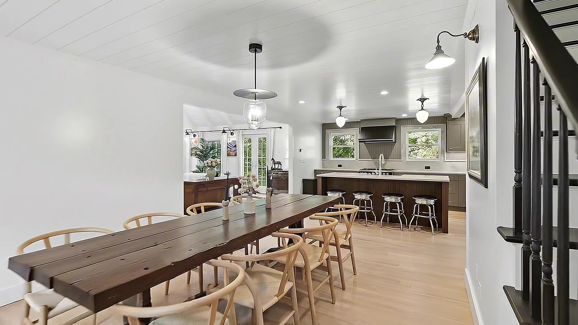 Open-concept dining area with long farmhouse wood table, wishbone dining chairs, glass pendant light, open to kitchen with island and bar stools, shiplap ceiling, and wrought-iron staircase