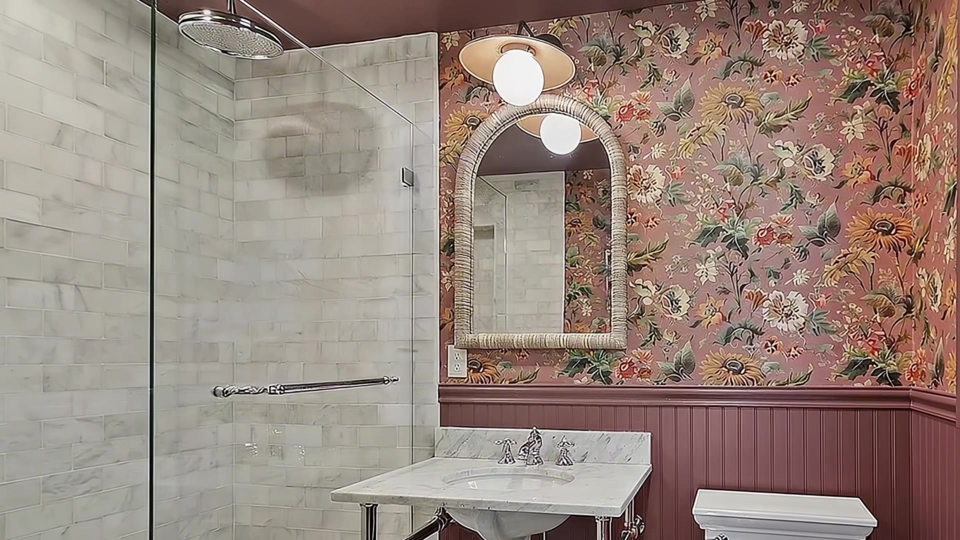Elegant bathroom with marble subway tile walk-in shower, rain showerhead, floral wallpaper, burgundy wainscoting, marble-topped vanity with vintage fixtures, and arched mirror