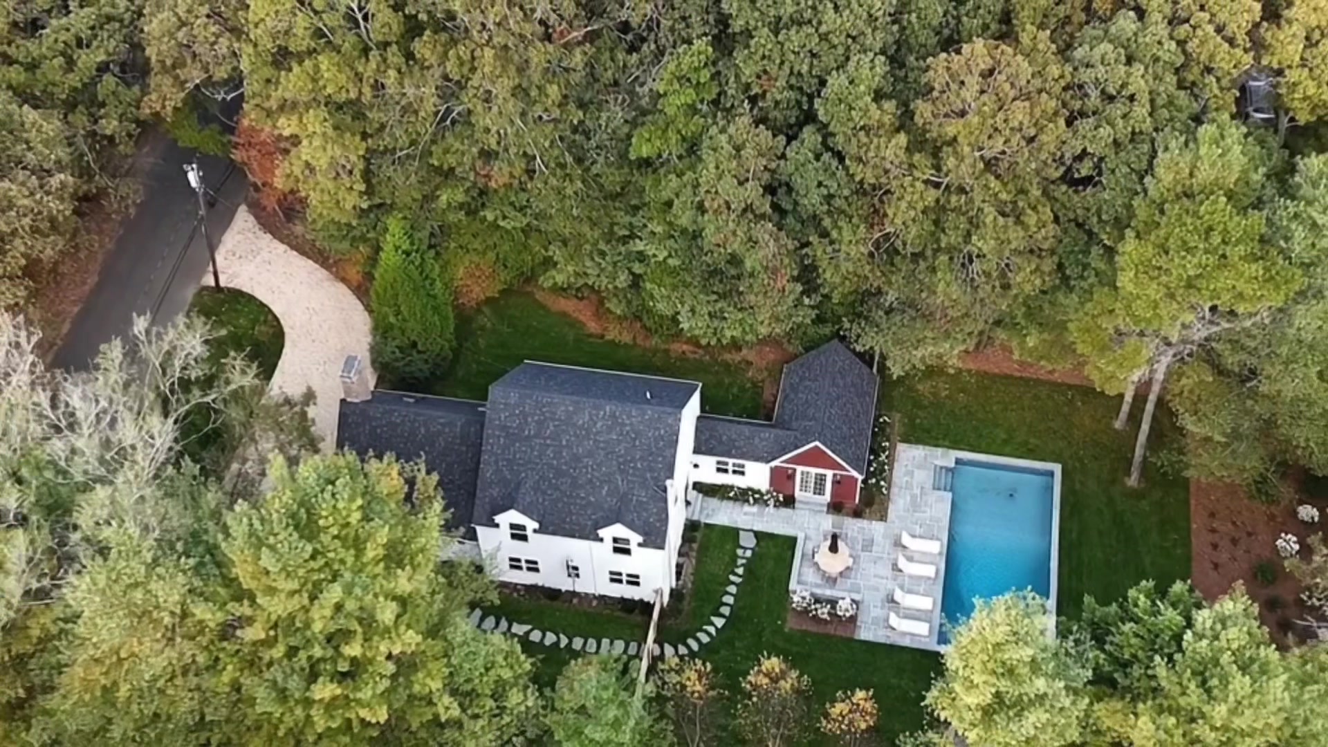 Aerial view of 6 East Gate Road showing the white shingled main house, red pool house, in-ground swimming pool, stone patio, circular driveway, and surrounding mature trees on nearly half an acre